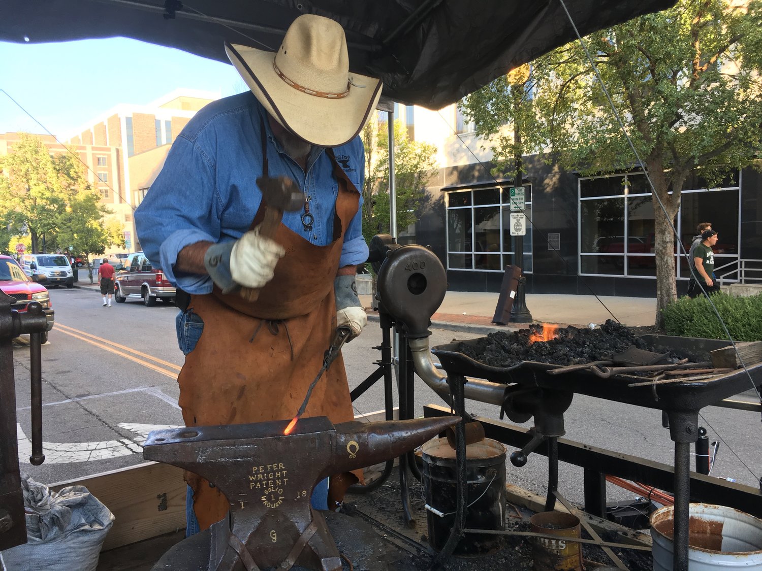 Blacksmith wearing a cowboy hat with hammer in hand working at a mobile forge giving a blacksmithing demonstration.