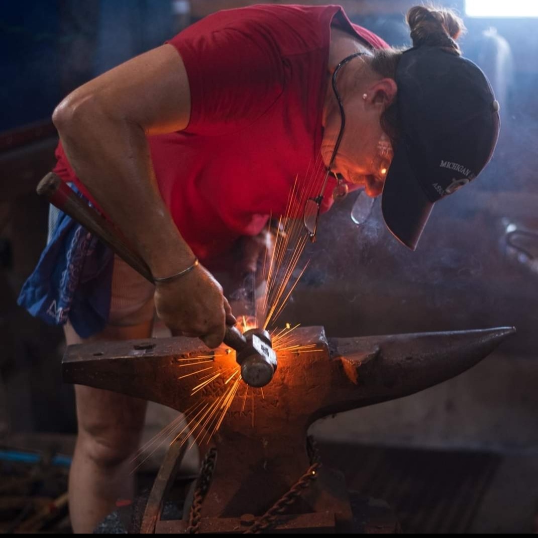 Woman in baseball cap bent over an anvil demonstrating a forging technique as sparks fly.